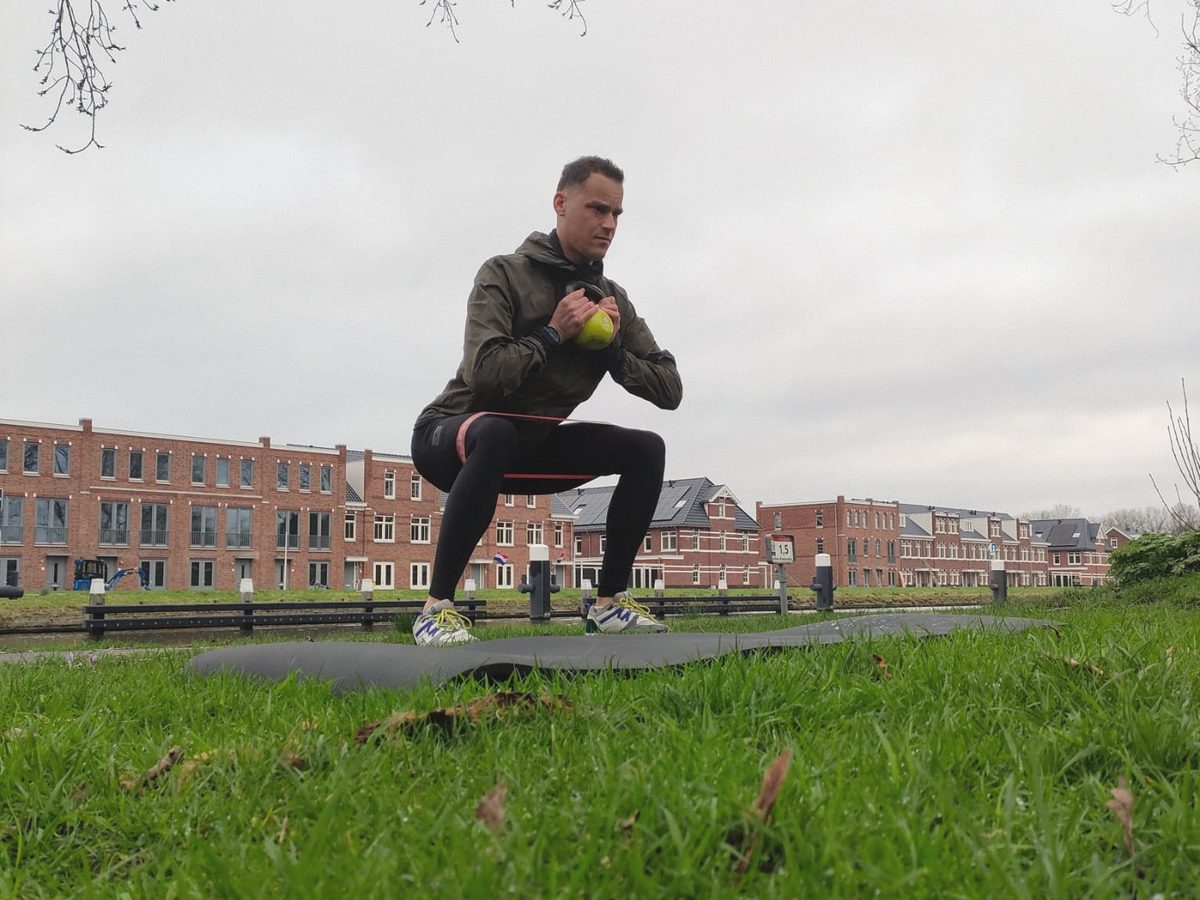 Iemand doet een kniebuiging met een kettlebell op een mat, met huizen op de achtergrond